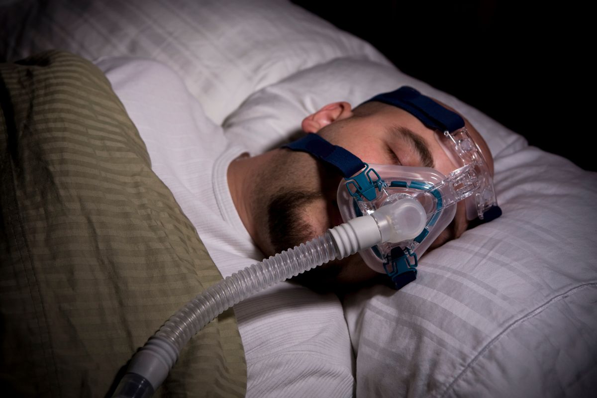 Image of a man sleeping with a CPAP machine, used to treat sleep apnea, emphasizing the risk factors for this condition. Sleep apnea is more common in older adults, men, and individuals who are overweight or obese. Other risk factors include smoking, alcohol use, and family history of sleep apnea. Understanding the risk factors for sleep apnea is important for identifying those at higher risk and promoting early diagnosis and treatment, which can improve overall health outcomes.